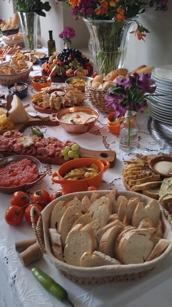 Mesa longa de entradas com cestas de pães, antepastos, tâbuas de frios e arranjos florais