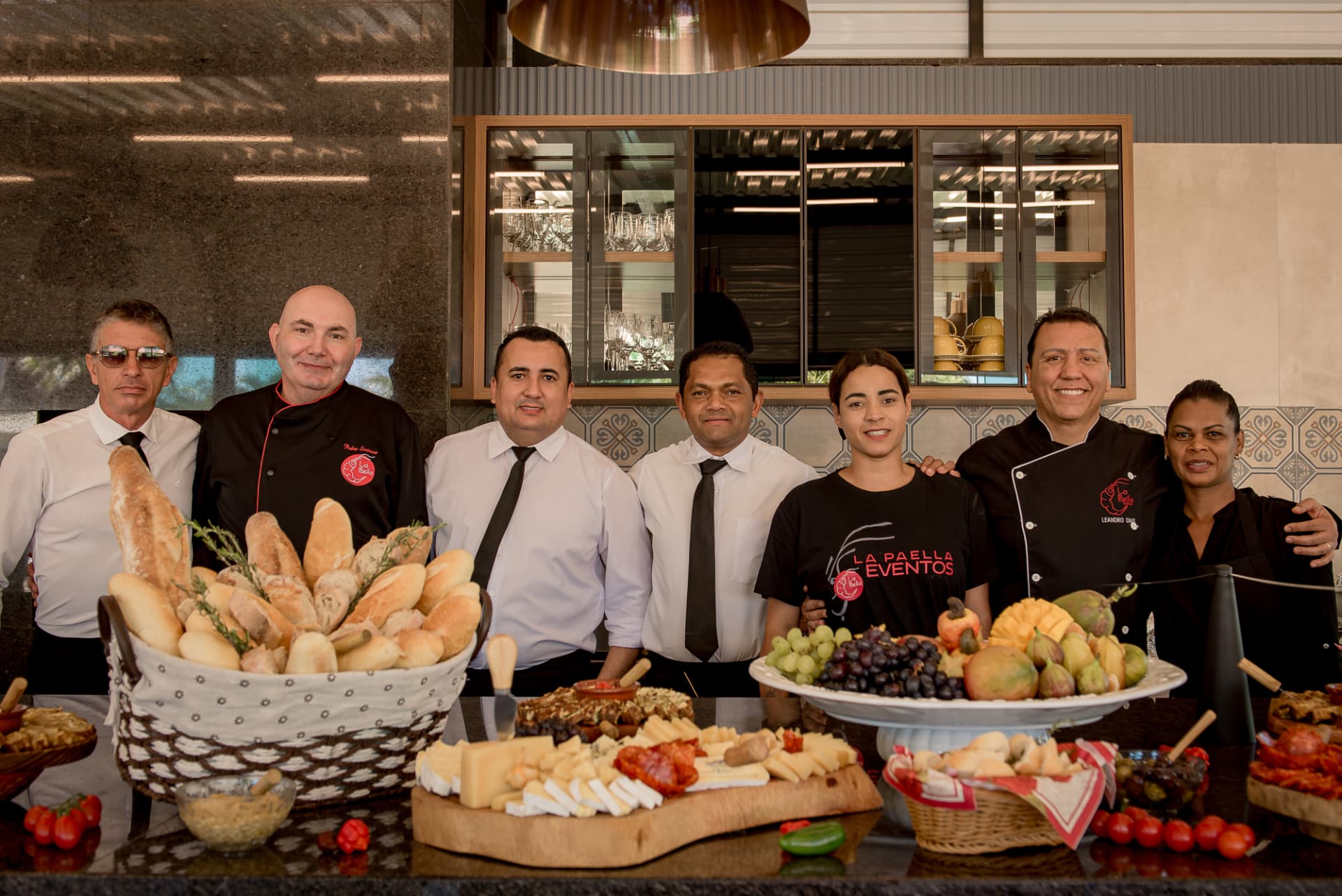 Equipe do La Paella Eventos reunida atrás de mesa com pães, queijos, frutas e antepastos