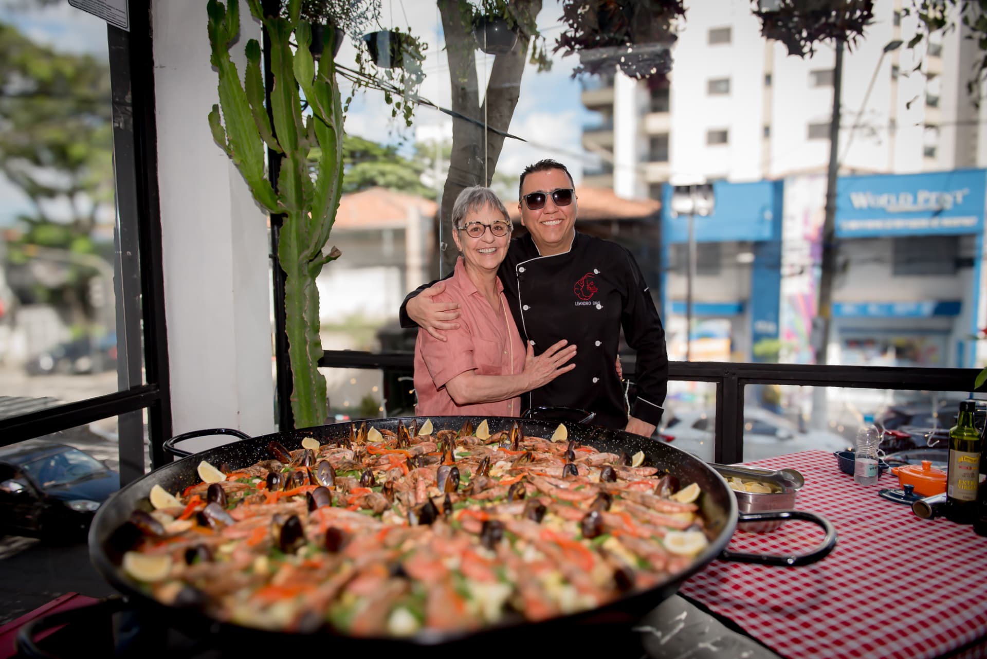 Chef Leandro Dias abraçado a cliente atrás da paella gigante de frutos do mar na varanda do restaurante