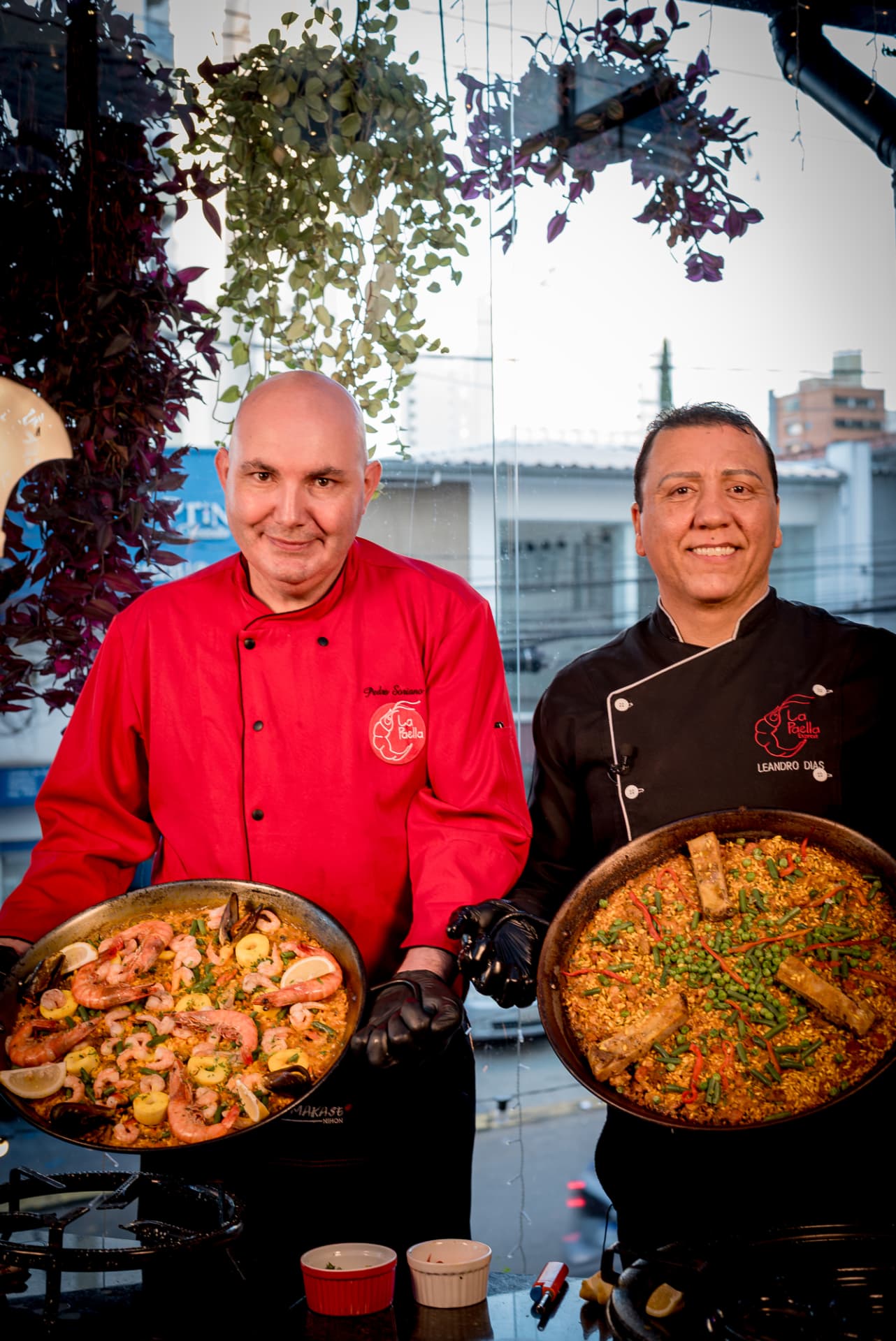 Leandro Dias e Pedro Soriano sorrindo, cada um segurando uma paellera pronta, em frente às janelas do restaurante